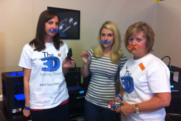 Team at The 3‑D Printing Store in Denver posing with colorful 3‑D‑printed mustaches and mini objects.