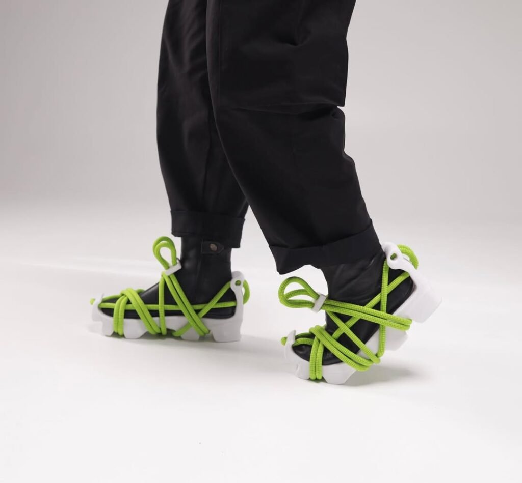 Close-up of UDRB sneakers worn with black pants, showing modular white 3D-printed soles and neon green climbing-rope laces wrapping around the foot.