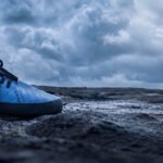 Close-up of a blue WAZP METIS 3D printed shoe worn outdoors on dark rocky ground under a cloudy sky.