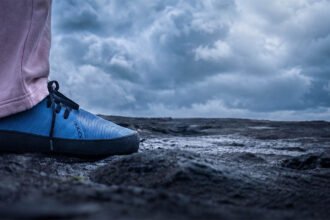 Close-up of a blue WAZP METIS 3D printed shoe worn outdoors on dark rocky ground under a cloudy sky.