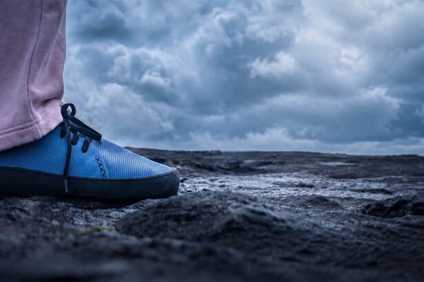 Close-up of a blue WAZP METIS 3D printed shoe worn outdoors on dark rocky ground under a cloudy sky.