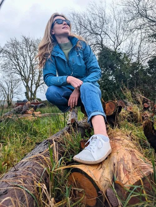 Person sitting on logs outdoors while wearing white WAZP METIS 3D printed shoes with jeans and a blue jacket.